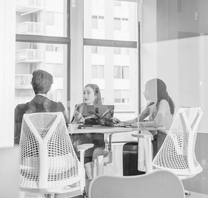 Three colleagues collaborate around a table with a laptop in a modern glass-walled office meeting room.