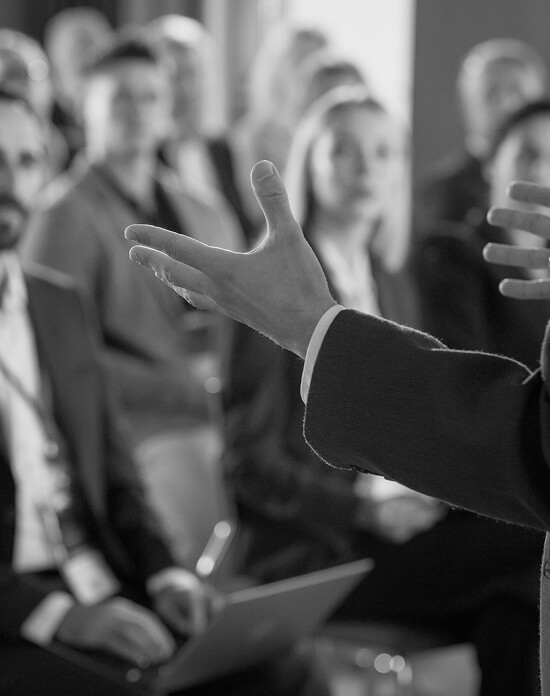 A speaker's outstretched hands gesture toward an attentive audience during a public affairs presentation in black and white.