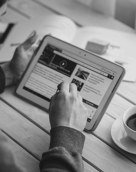 A person browsing a news website on a tablet at a wooden table with a coffee cup nearby, in black and white.
