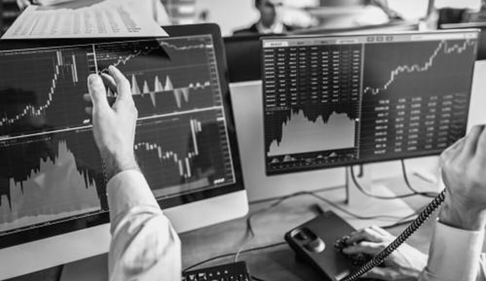 A financial professional analyzing stock market charts and trading data on dual monitors at a desk.