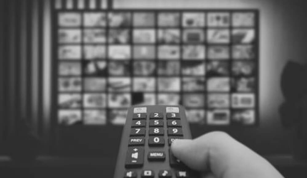 A hand pointing a remote control at a television screen displaying a grid of entertainment content thumbnails.