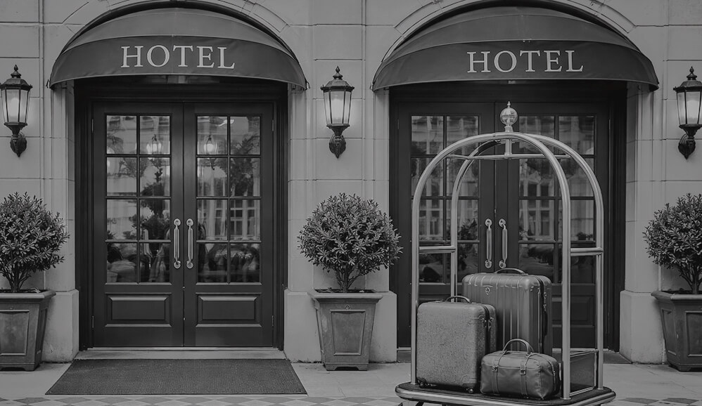 Elegant hotel entrance with arched awnings, glass doors, lantern sconces, and a luggage cart with suitcases.