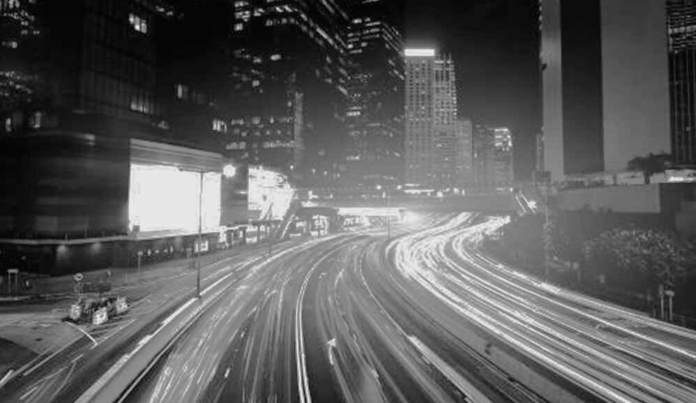 Long-exposure black-and-white photograph of a busy multi-lane highway curving through a city skyline at night.