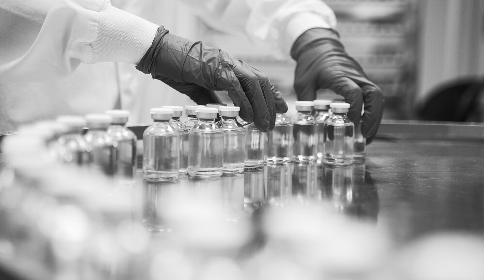 Gloved hands in lab coats arrange sealed pharmaceutical vials on a reflective surface in a production facility.