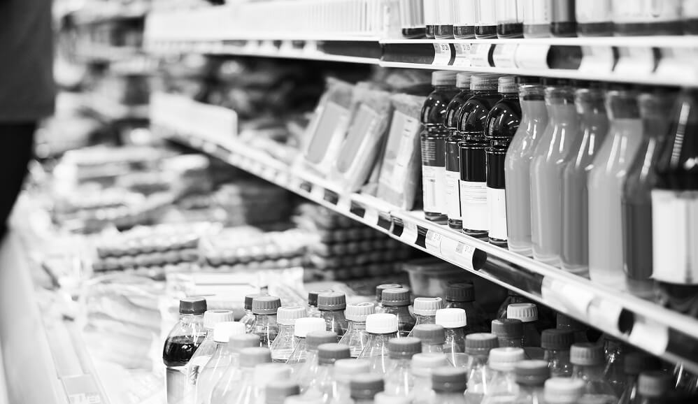 Black-and-white close-up of grocery store shelves stocked with various bottled beverages and packaged food products.