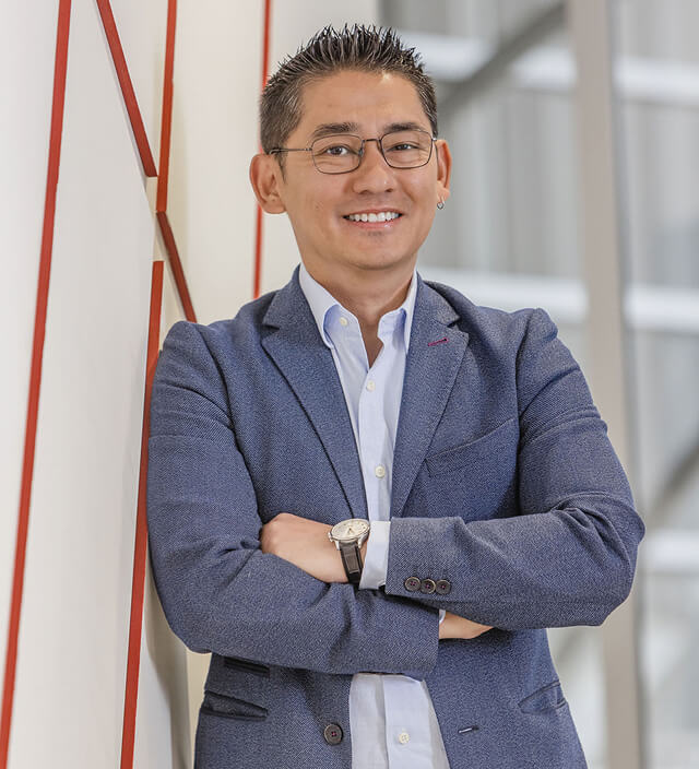 Andrew de Silva smiling with arms crossed, wearing glasses, a blue blazer, and a white open-collar shirt in a modern office