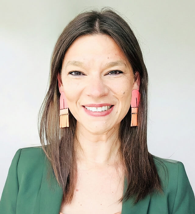 Anna Blender smiling in a professional headshot, wearing a green blazer and bold coral statement earrings.