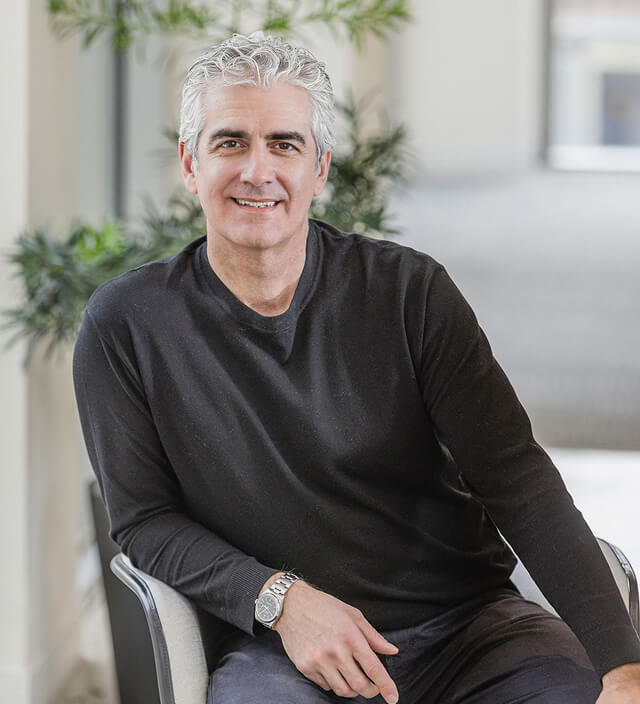 Anthony Michelini smiling in a black sweater, seated in a modern office setting with green plants behind him.
