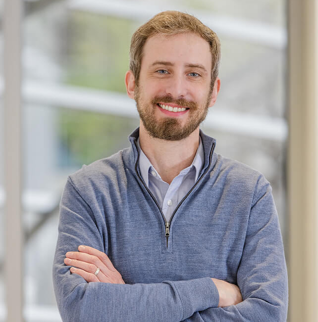 Eric Zimmermann smiling with arms crossed, wearing a blue quarter-zip sweater over a collared shirt, with a blurred office