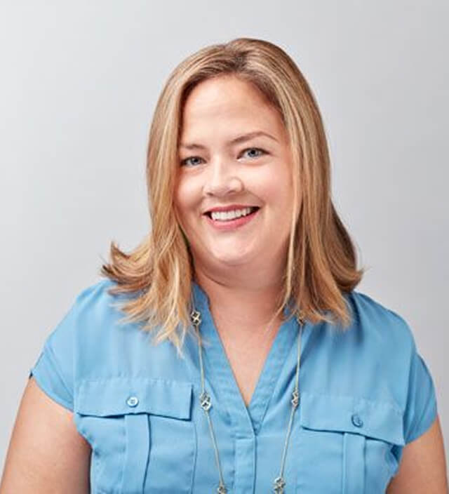 Professional headshot of Sara Gormley smiling, wearing a blue blouse and a long necklace against a gray background.